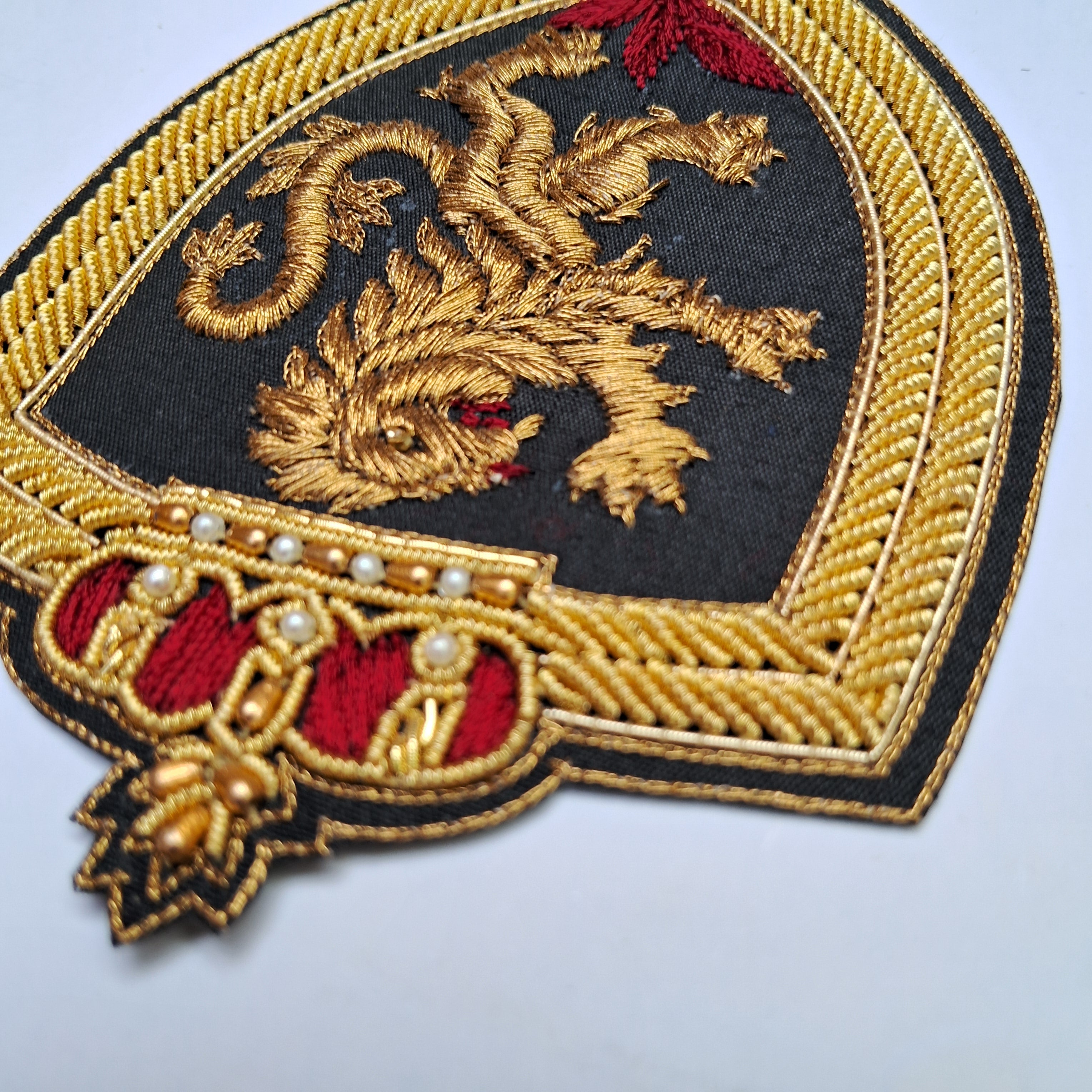An embroidered heraldic patch featuring a gold lion rampant on a black shield, bordered by gold rope trim and topped with a red crown motif with small pearl-like beads, shown on a light gray background.
