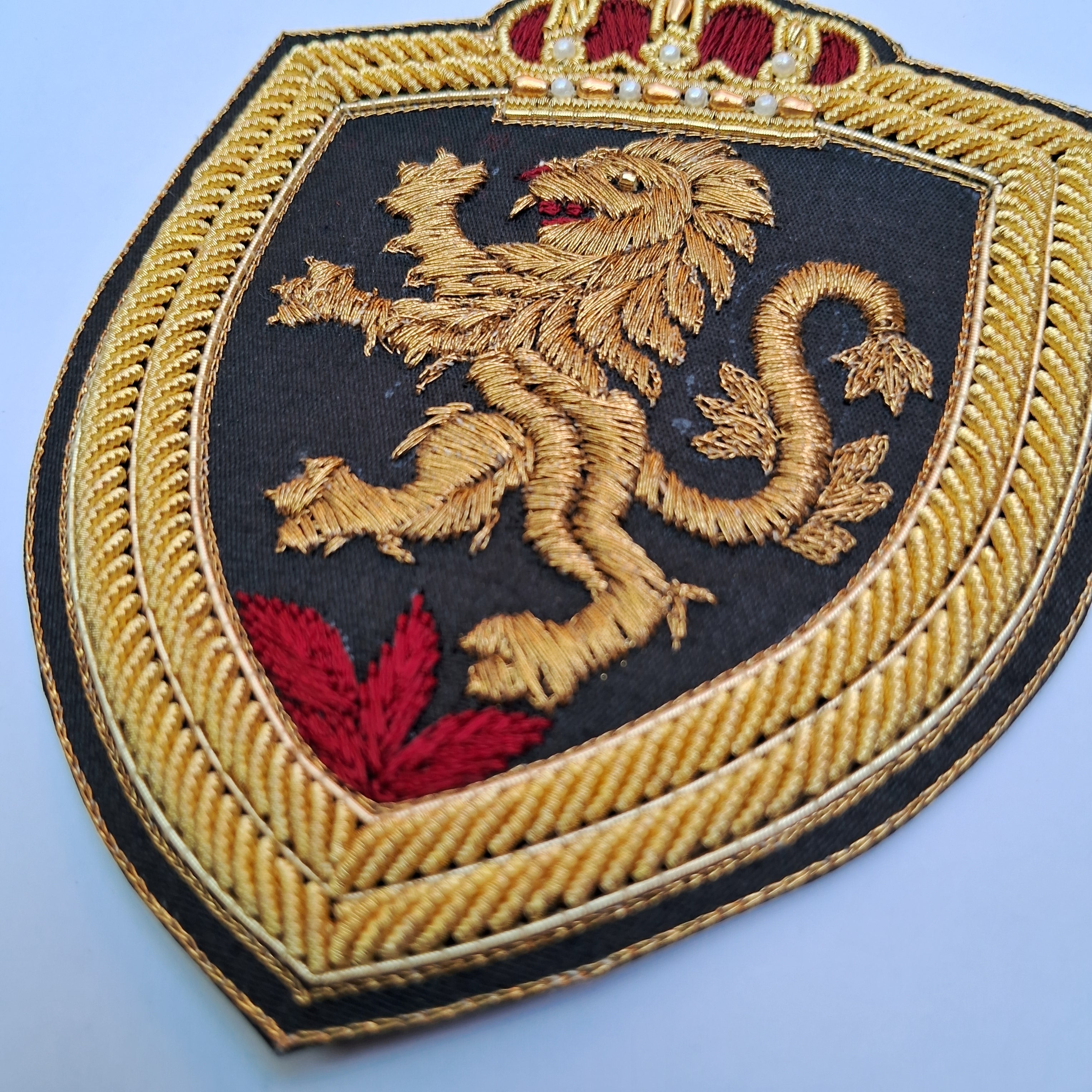 An embroidered shield-shaped patch featuring a gold lion rampant on a black background, bordered by gold rope embroidery with red crown and leaf details, photographed on a light blue background.