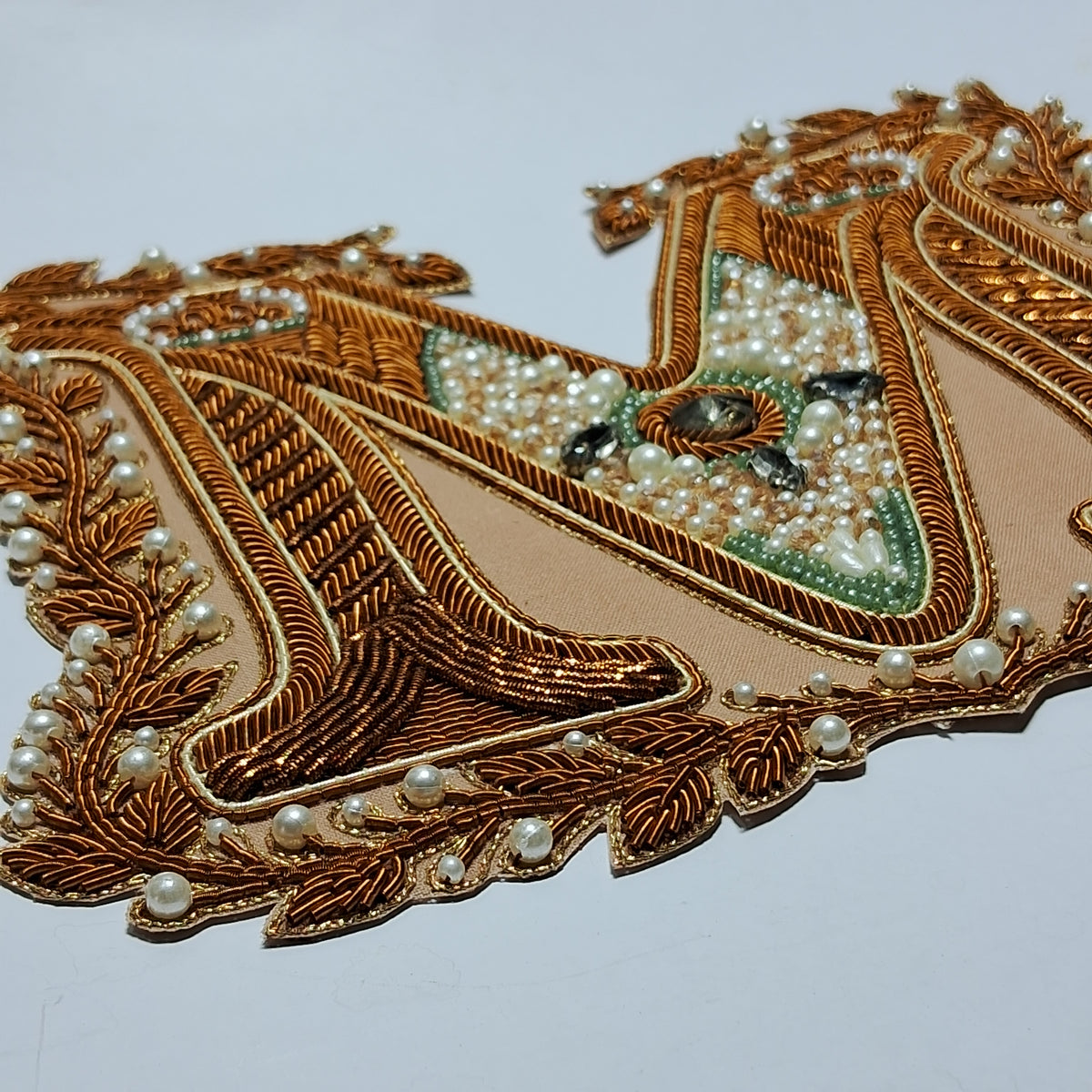 An ornate embroidered patch featuring metallic gold threadwork, white faux pearls, and a central green and gold pattern on a light beige background, set against a plain white backdrop.