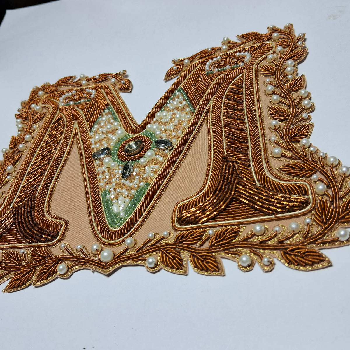 An embroidered letter 'M' applique patch featuring intricate gold metallic embroidery, white pearl beads, and green accents on a beige background, shown on a plain white surface.