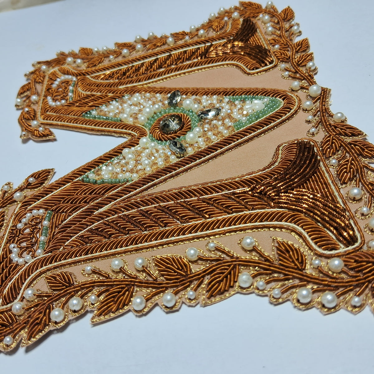 An ornate embroidered letter 'A' applique patch in metallic bronze and gold thread, embellished with white faux pearls, green beadwork, and a central dark jewel, shown on a plain white background.