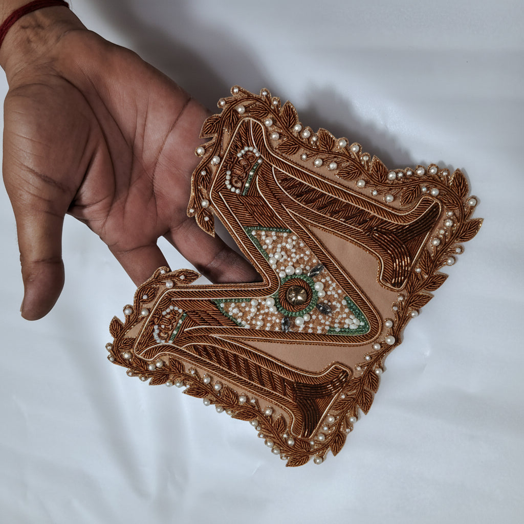 A hand holding a decorative embroidered patch or applique with intricate brown beadwork, fringe trim, and a central floral motif on a white fabric background.