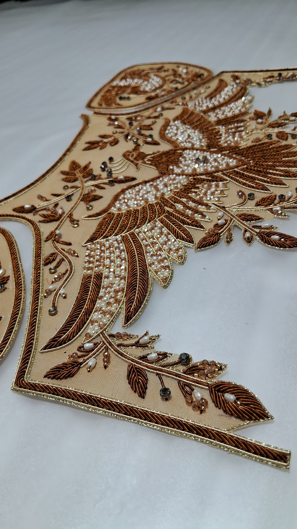A close-up of an ornate beige embroidered fabric piece featuring a bird motif with brown threadwork, white pearl-like beads, and small dark embellishments on a light background.