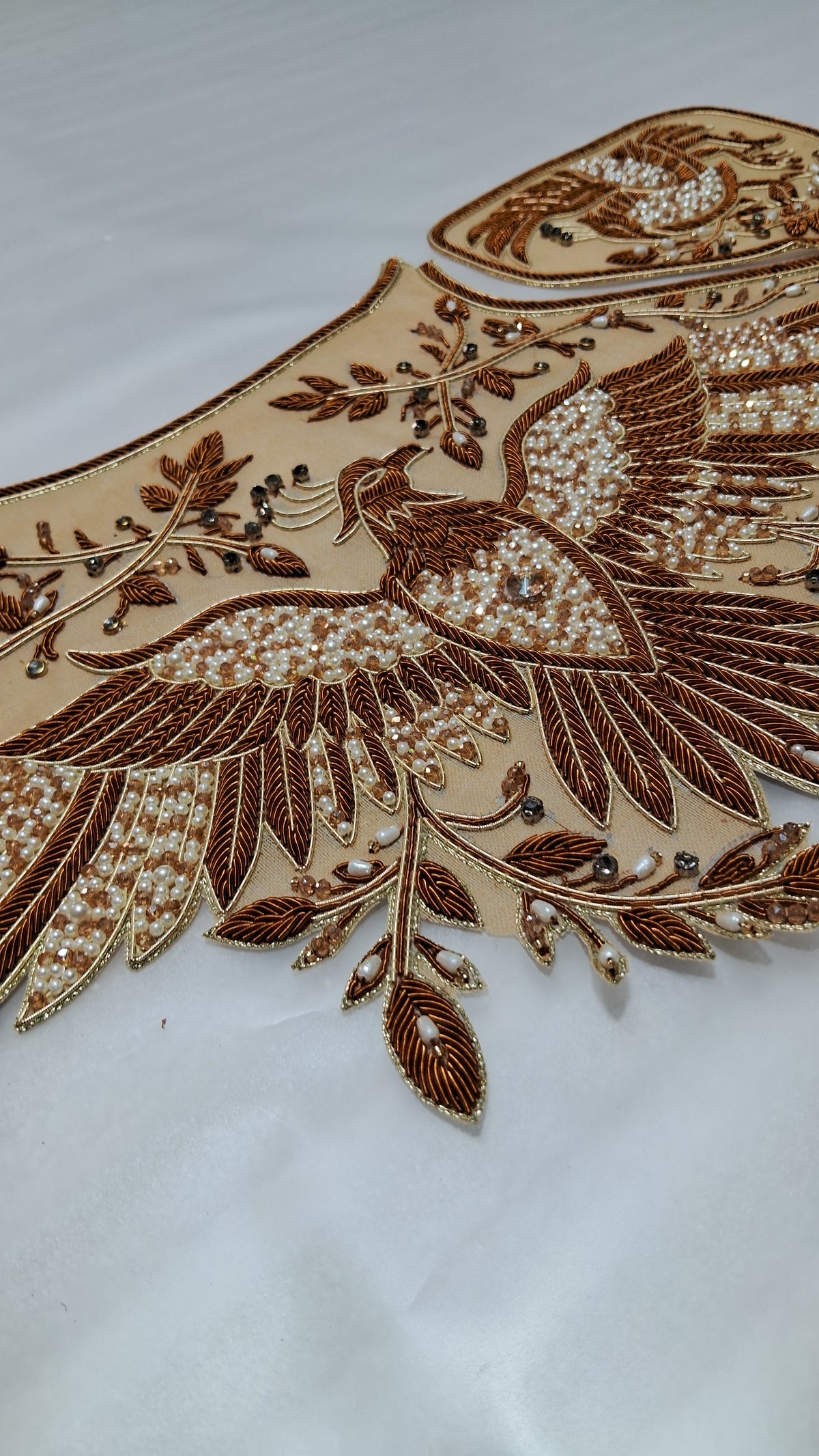 A close-up of a beige embroidered applique featuring a detailed brown bird with white pearl-like beadwork and leafy branches, laid on a white fabric background.