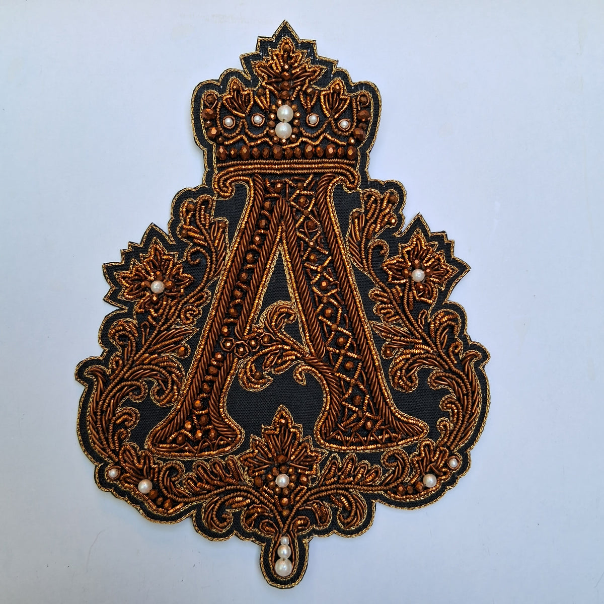 An ornate embroidered monogram patch featuring a large letter 'A' with a crown motif, surrounded by floral scrollwork and small pearl-like beads on a black fabric background, shown against a light blue background.