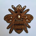 An ornate embroidered bee applique patch featuring intricate metallic gold and bronze beadwork, black outlines, and small white pearl accents on a light gray background.
