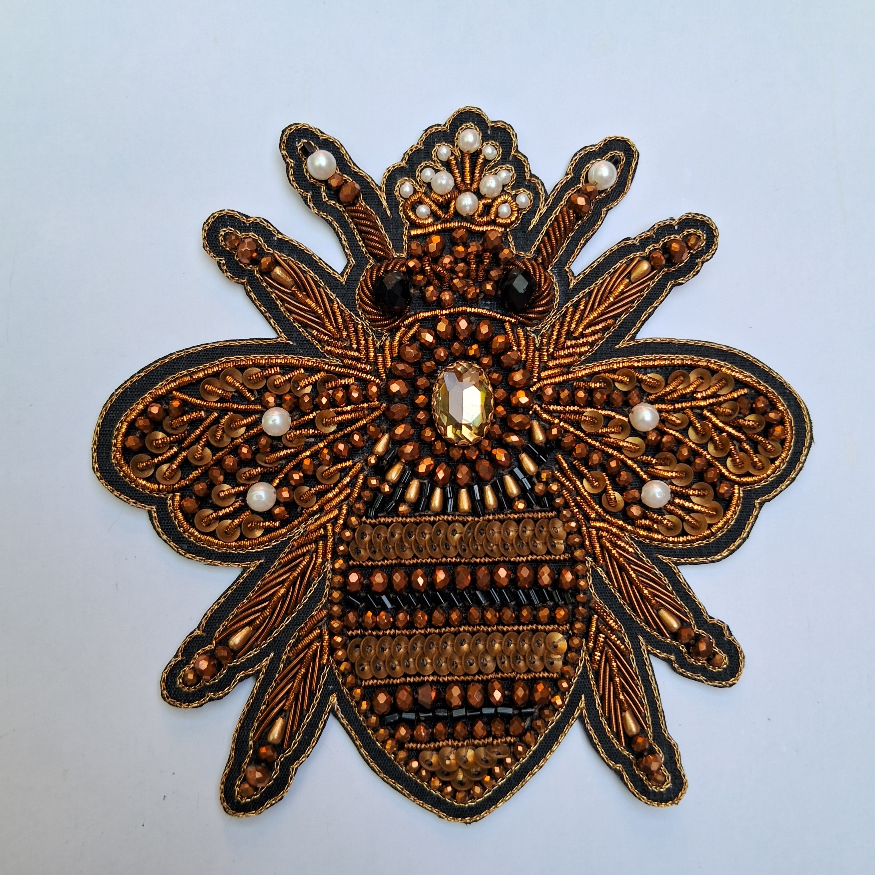 An ornate embroidered bee applique patch featuring intricate metallic gold and bronze beadwork, black outlines, and small white pearl accents on a light gray background.