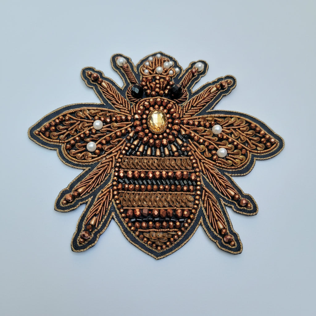 A close-up of a decorative bee applique or patch made with copper-toned beads, brown sequins, black beads, white pearl-like accents, and a faceted clear center jewel on a light blue background.