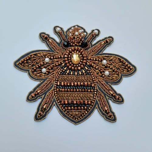 A close-up of a decorative bee applique or patch made with copper-toned beads, brown sequins, black beads, white pearl-like accents, and a faceted clear center jewel on a light blue background.
