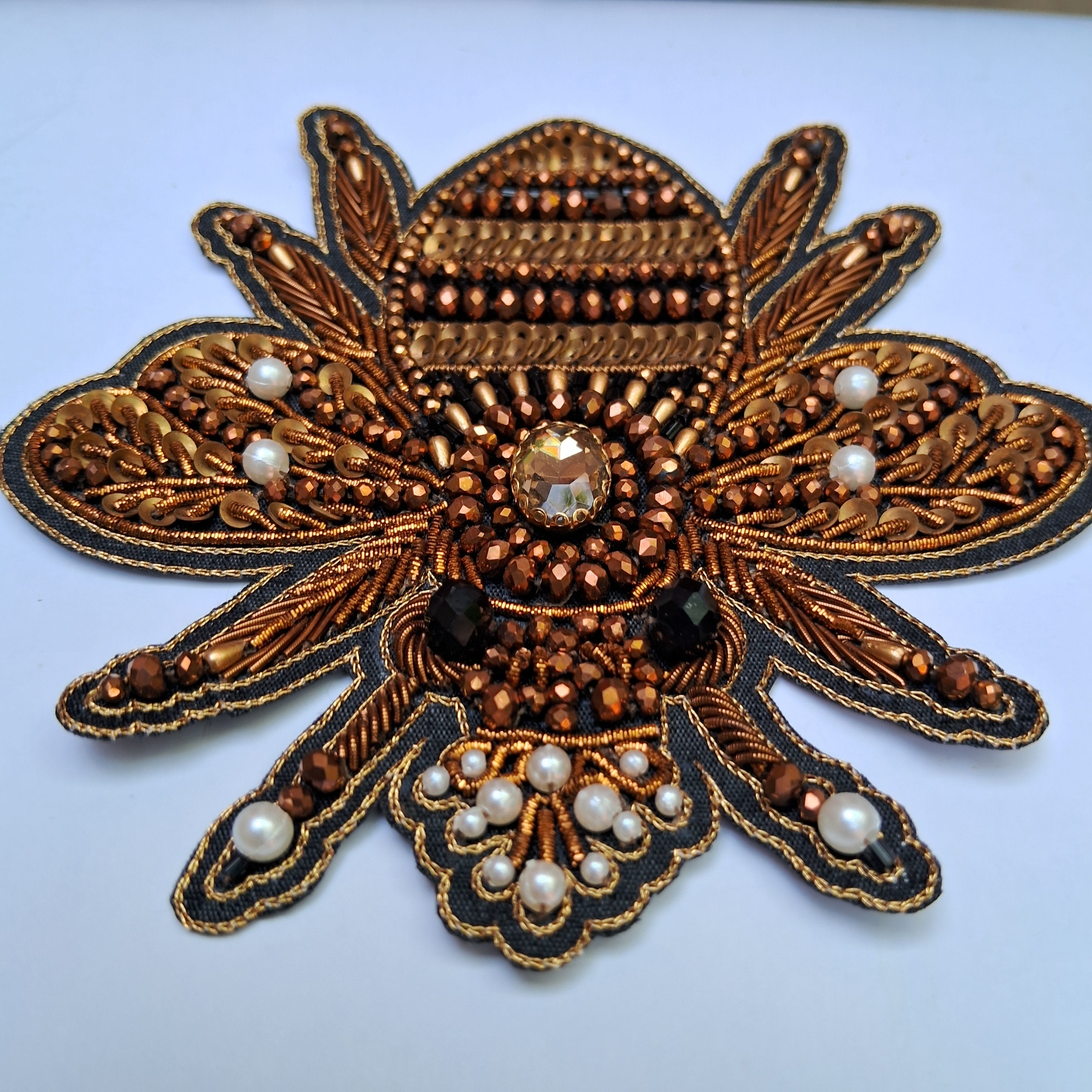 An ornate embroidered applique patch shaped like a stylized flower or sunburst, featuring metallic bronze and gold threadwork, faux pearls, and rhinestones on a light blue background.