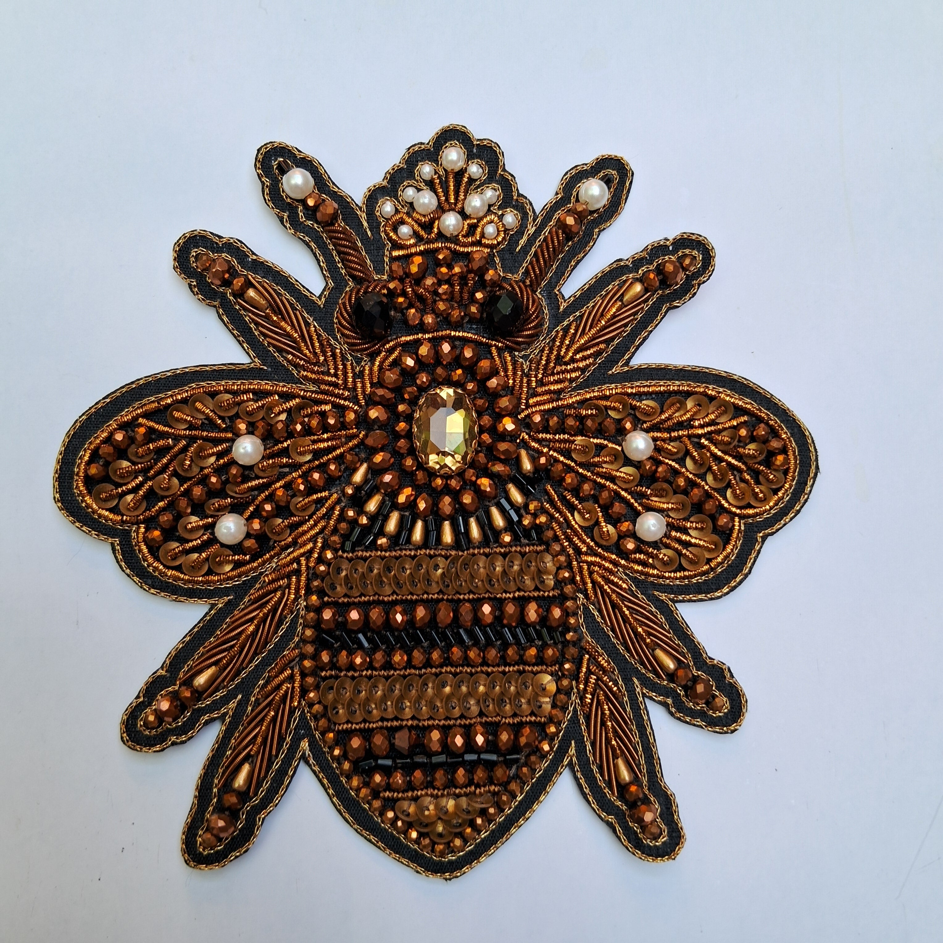 An ornate embroidered bee applique patch with metallic bronze and gold threadwork, black outlines, and white pearl-like bead accents on a light gray background.