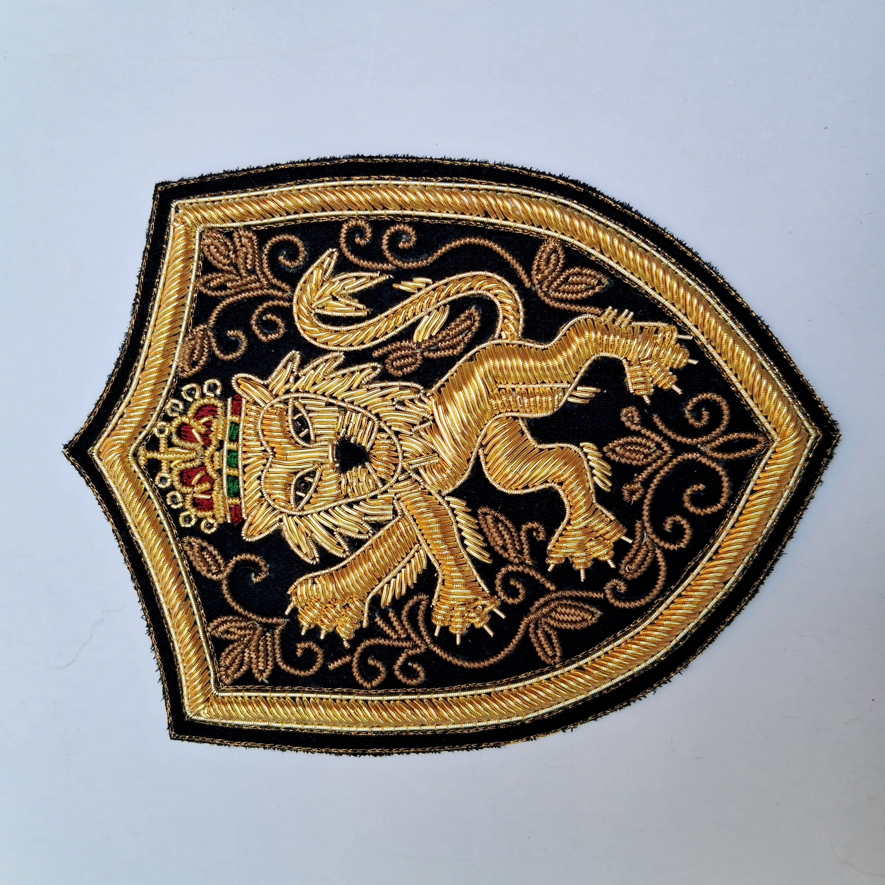 An embroidered shield-shaped patch featuring a golden lion rampant on a black background with ornate gold scrollwork and a small red and green crown detail, shown on a plain light background.