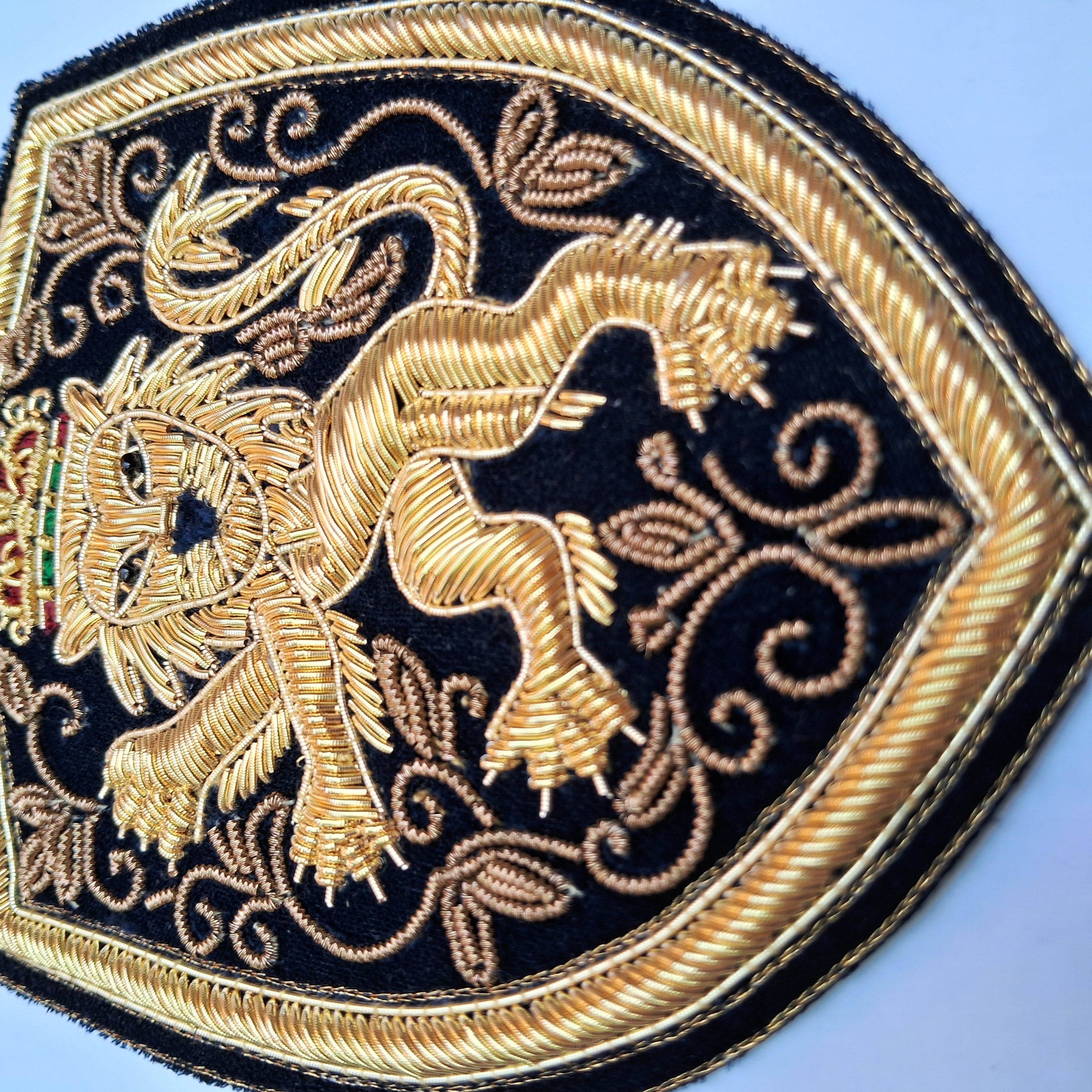 Close-up of an embroidered shield-shaped patch featuring a golden lion and ornate scrollwork on a black background, with a twisted gold border.