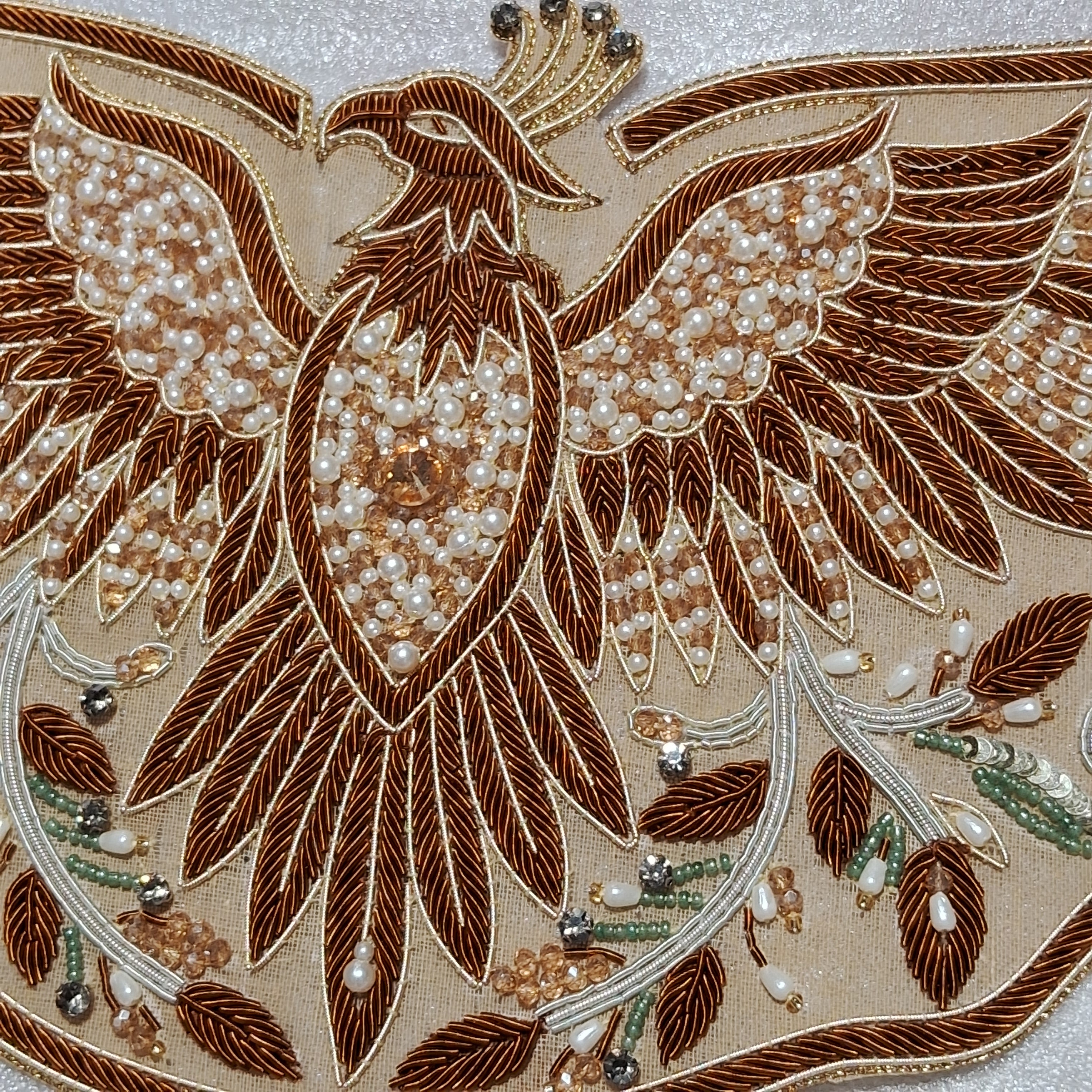 Close-up of an ornate embroidered phoenix design with brown thread, white pearl-like beads, and small green and silver decorative accents on a light beige fabric background.