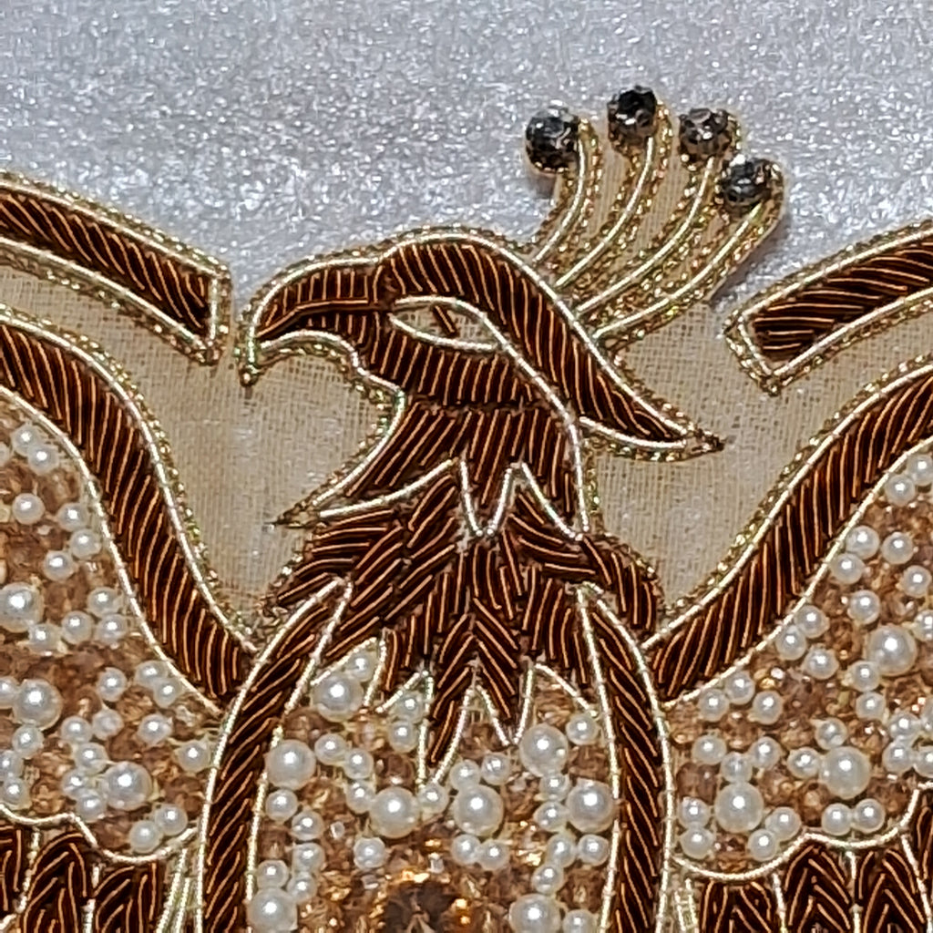 Close-up of an ornate embroidered phoenix design on a garment, featuring metallic gold and bronze threadwork, white pearl beads, and dark rhinestone accents on a textured light gray fabric background.