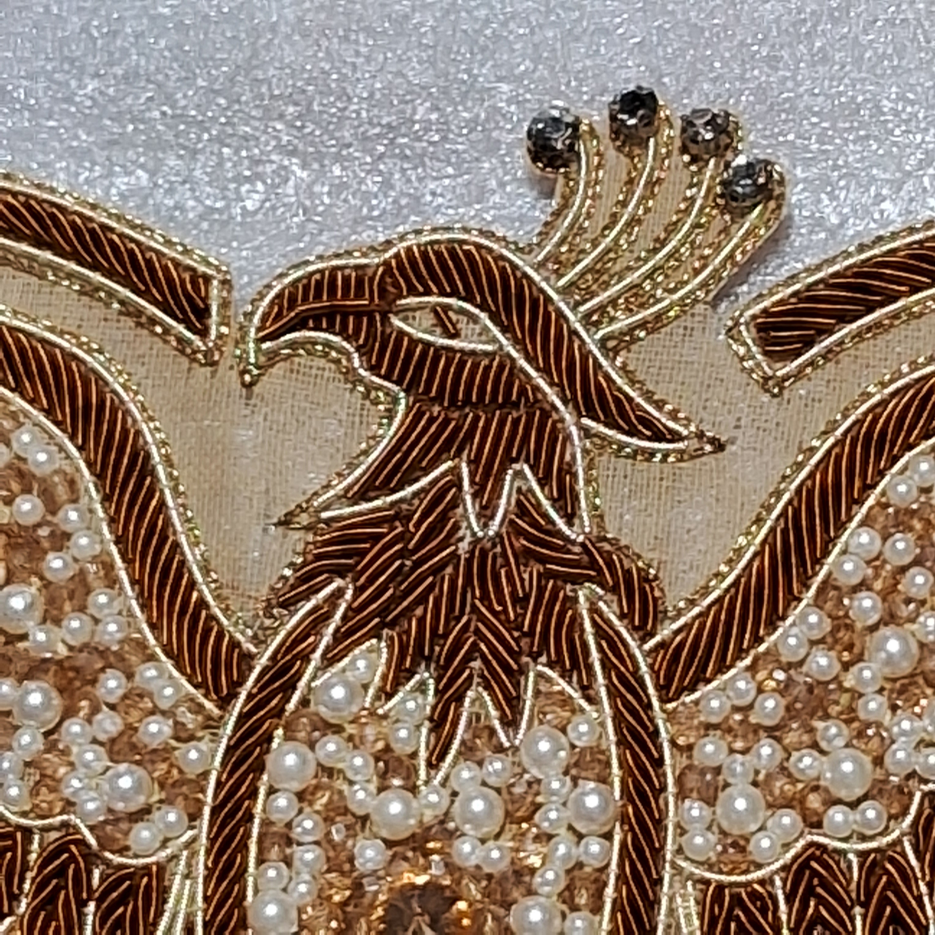Close-up of an ornate embroidered phoenix design on a garment, featuring metallic gold and bronze threadwork, white pearl beads, and dark rhinestone accents on a textured light gray fabric background.