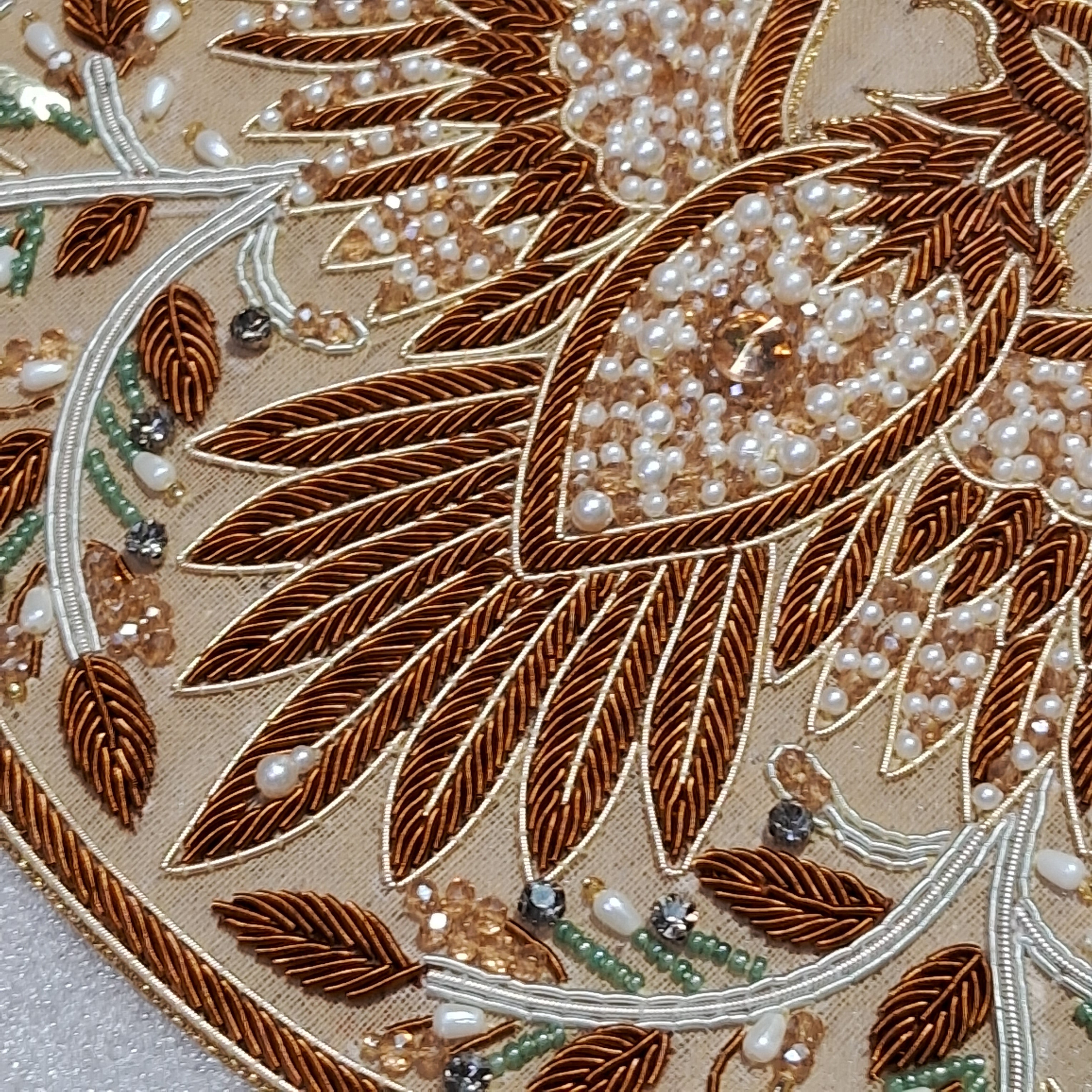 Close-up of ornate embroidered fabric featuring large brown leaf motifs, white pearl-like beads, and metallic gold and silver thread details on a beige textile background.