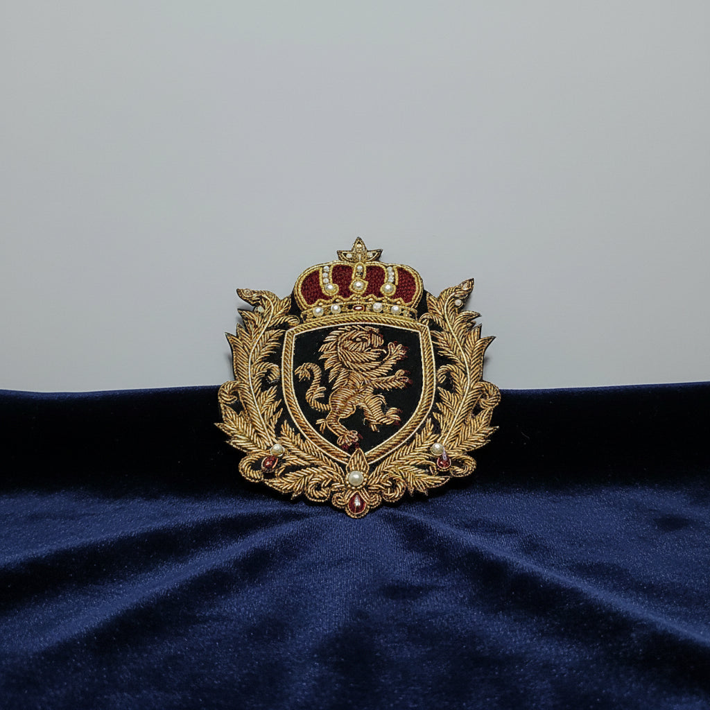 An ornate embroidered crest patch featuring a gold lion rampant on a black shield, topped with a jeweled crown and surrounded by a gold leafy wreath on a light blue background.