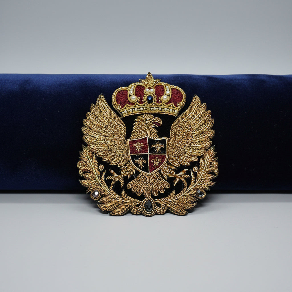 An embroidered double-headed eagle crest patch with a crown, gold thread wings, and a red and black shield, shown on a plain light background.