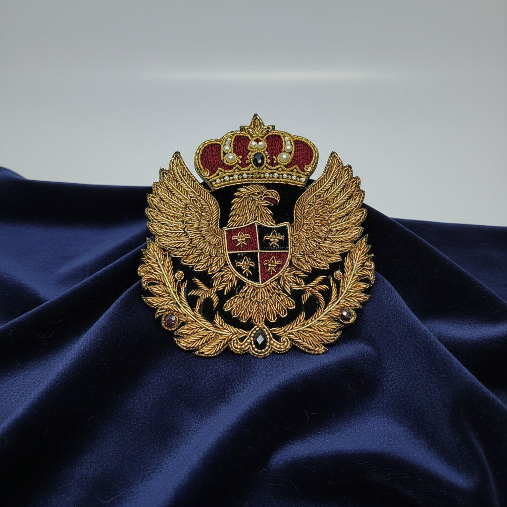 An embroidered double-headed eagle crest patch with a crown, gold thread wings, and a red and black shield, shown on a plain light background.