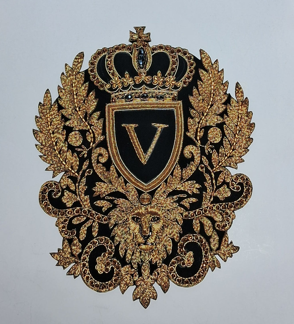An ornate embroidered crest patch featuring a gold lion, crown, and laurel wreath on a black background with a central shield containing a gold letter V, shown against a plain light gray background.