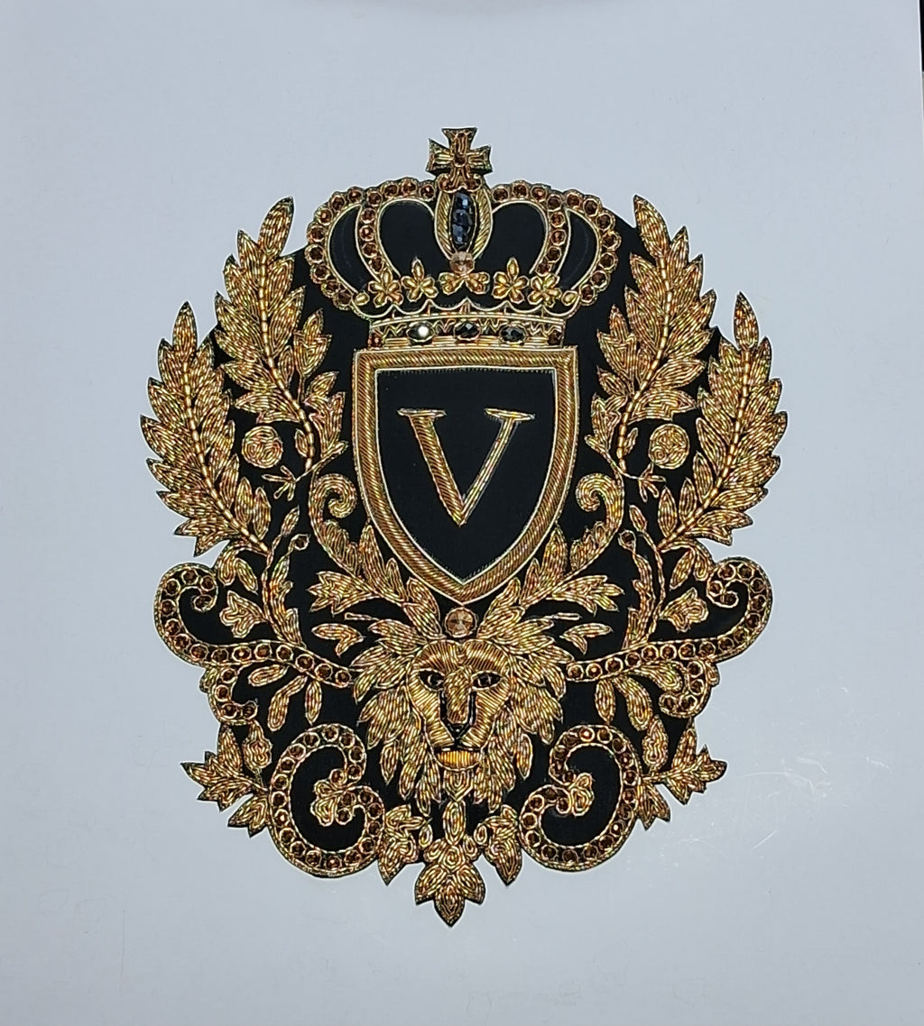 An ornate black and gold embroidered decorative emblem featuring a crown, shield with a large letter V, laurel leaves, a lion head, and scrollwork on a plain light background.