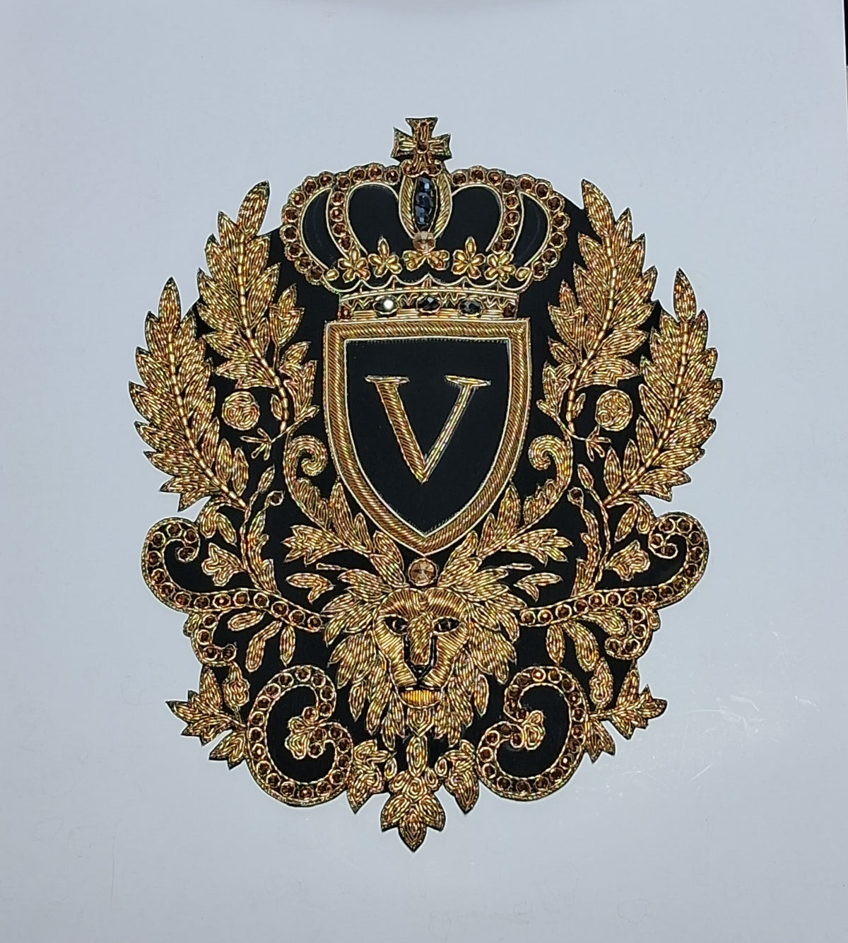 An ornate black and gold embroidered decorative emblem featuring a crown, shield with a large letter V, laurel leaves, a lion head, and scrollwork on a plain light background.