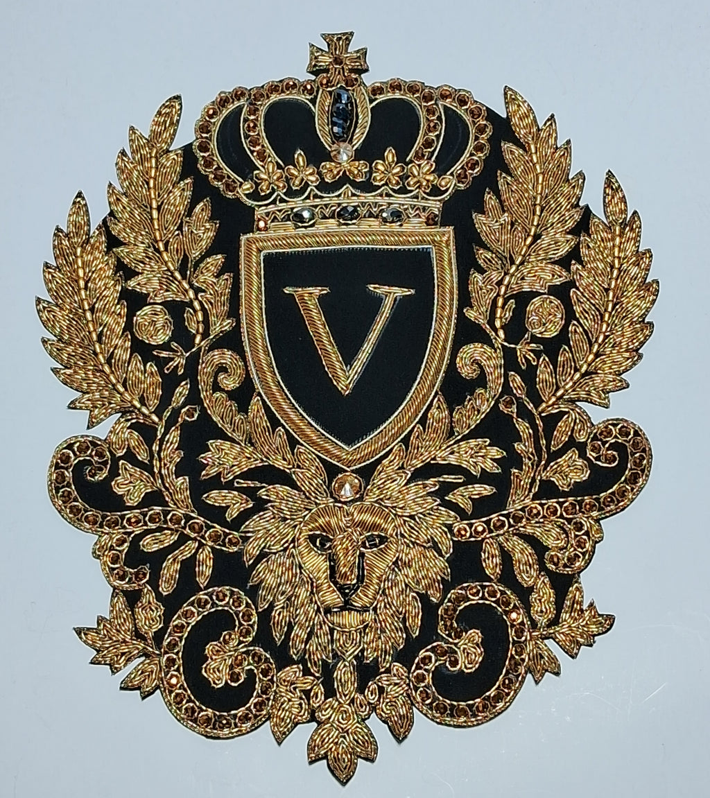 An ornate embroidered crest patch featuring a black background with intricate gold embroidery, including a crown, shield with a large letter V, laurel leaves, a lion head, and scrollwork.