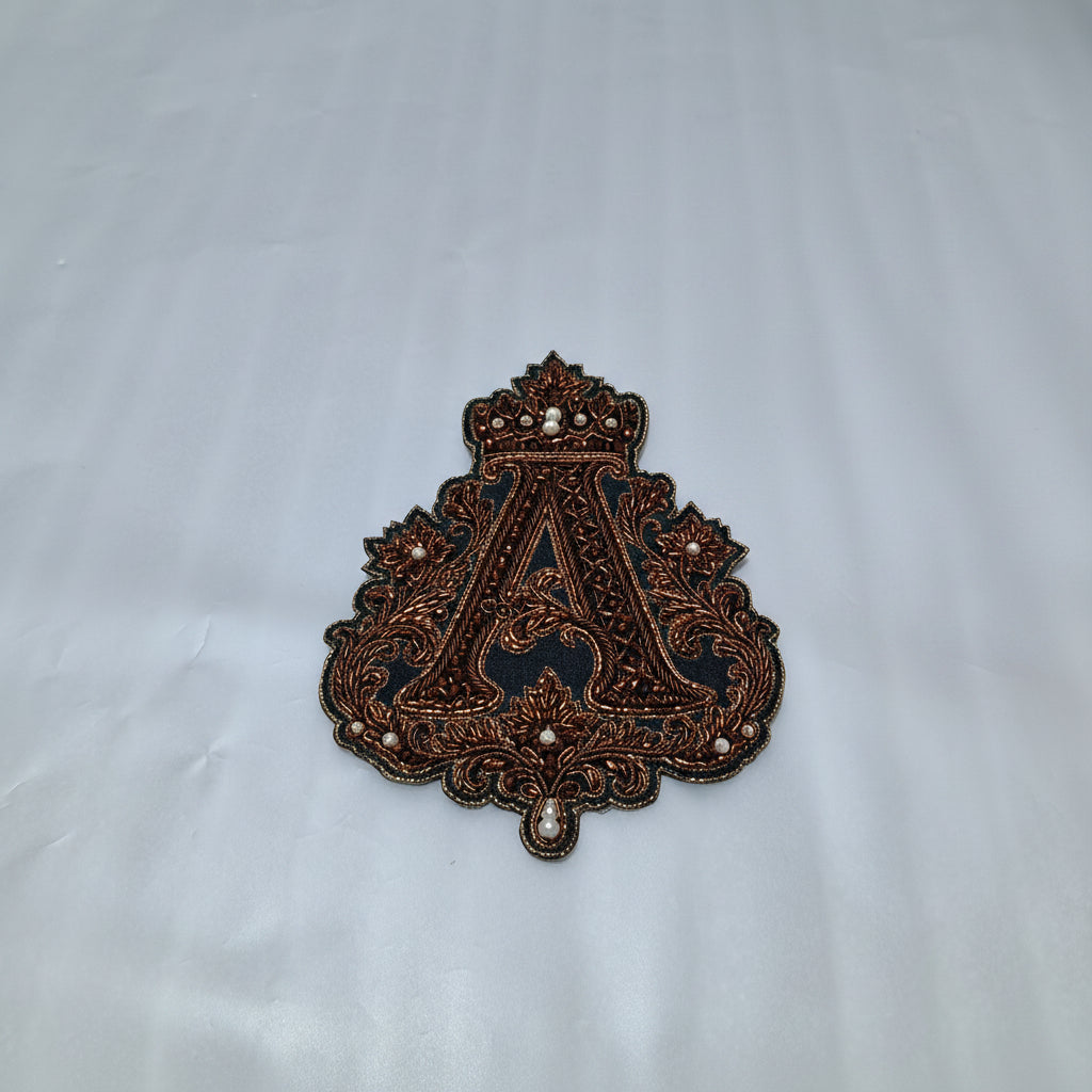 An ornate embroidered patch featuring a large letter 'A' with a crown motif, held in a person's hand against a white fabric background.