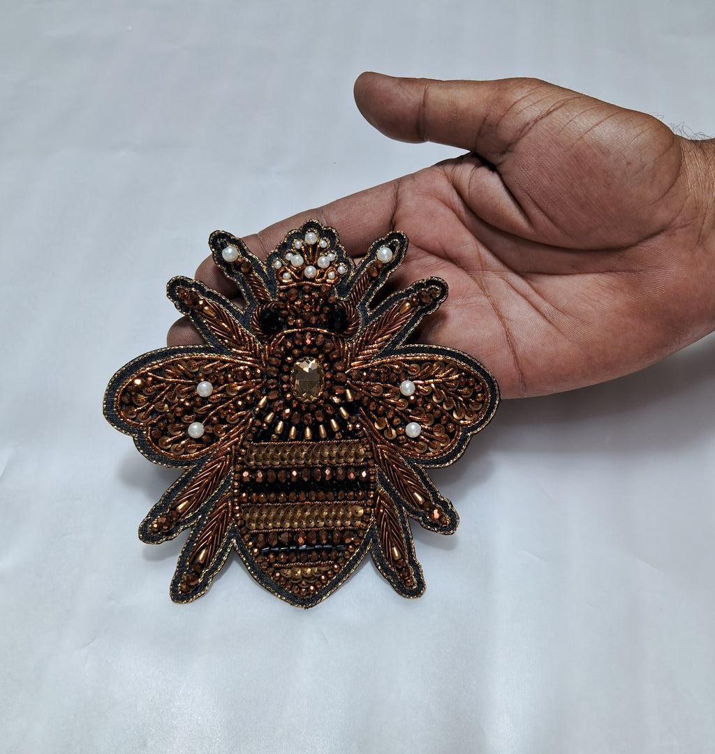 An ornate embroidered bee applique or brooch in metallic bronze, brown, and gold tones with small pearl-like white accents, shown resting on a white fabric surface with a person's hand holding it for scale.
